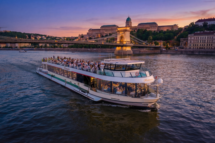 River cruise ship on water at sunset with historic bridge and buildings in background.