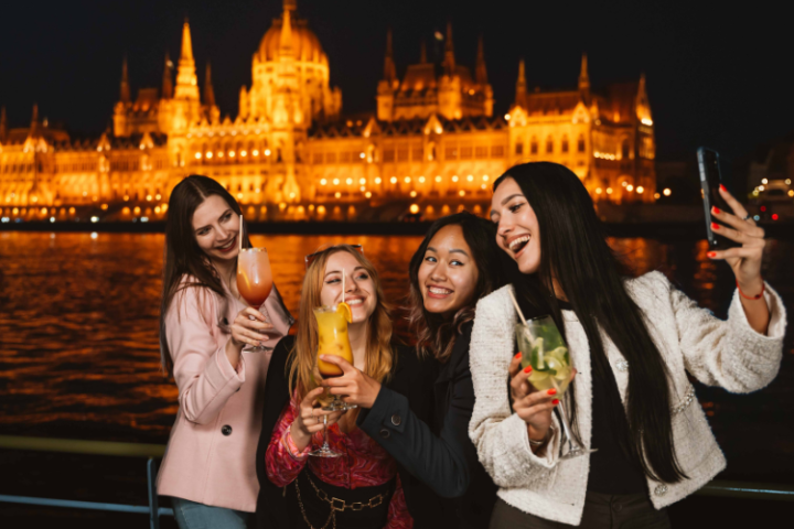 Four people taking a selfie with drinks at night, illuminated building in the background.