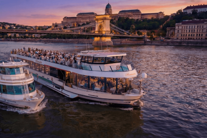 Two boats on a river at sunset with a lit bridge and cityscape backdrop.