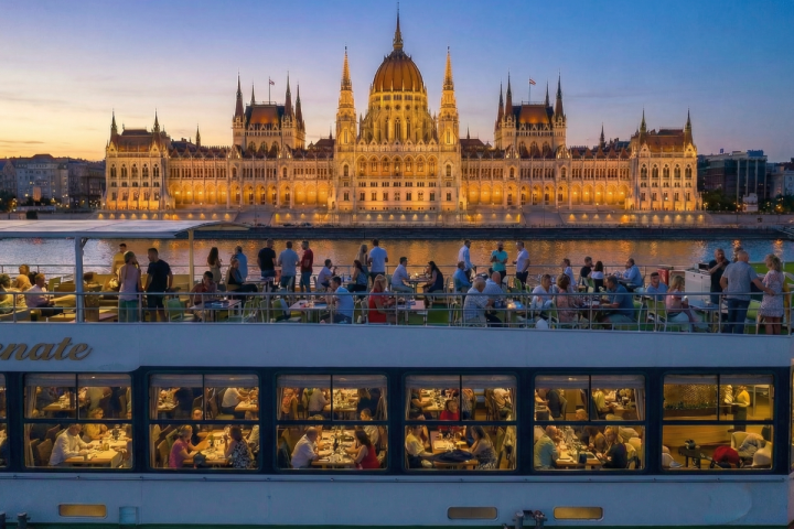 People dining on a boat with a lit building in the background at sunset.
