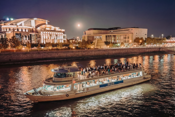 Lit cruise boat on river at night with city buildings and full moon in background.