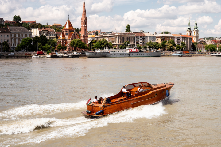 A wooden motorboat on a river with a cityscape and church spires in the background.
