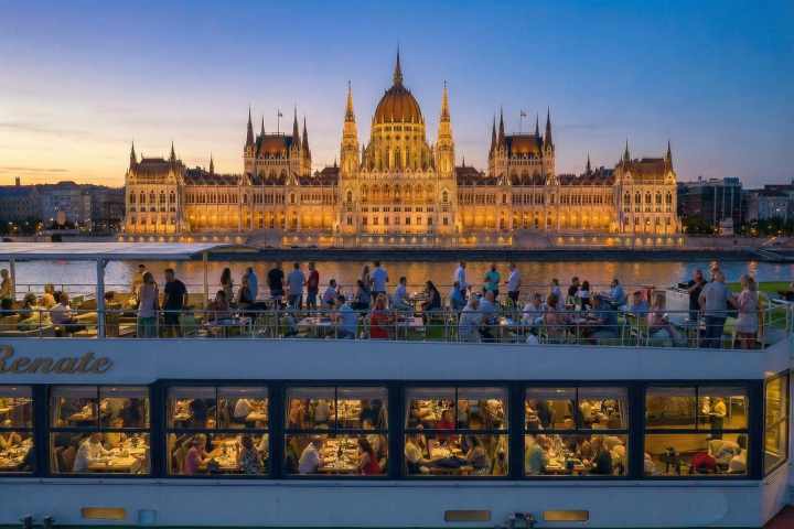 People dine on a cruise ship at dusk, with an illuminated parliament building in the background.