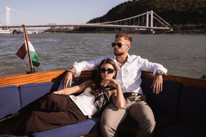 Couple relaxing on a boat with a river and bridge in the background.