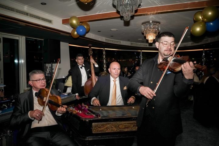 Musicians in formal wear play violins and bass at a decorated event with chandeliers and balloons.