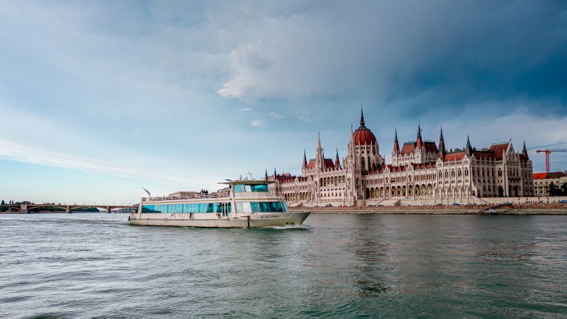 Boat on river with large historic building, cloudy sky in the background.