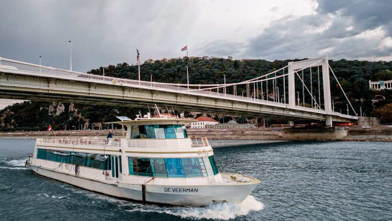 Tour boat on a river beneath a bridge, with cloudy sky and hills in background.