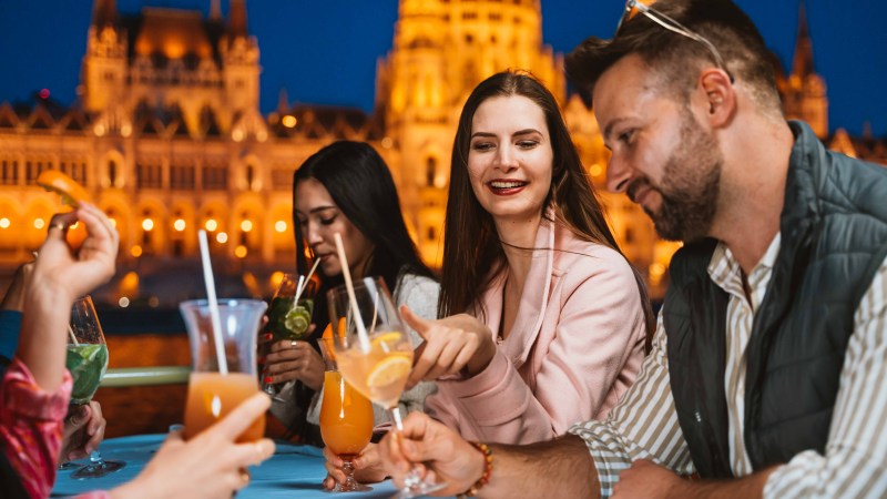 Group of friends enjoying cocktails at an outdoor evening gathering near a lit-up historic building.