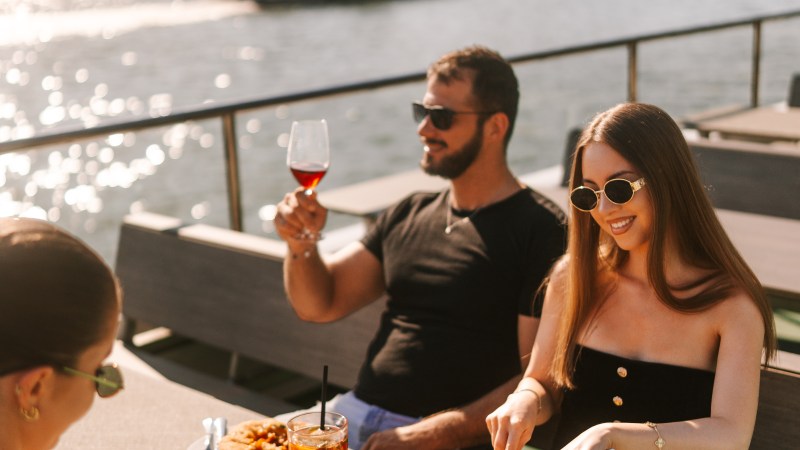 Three people dining outdoors by a river, with two women and a man enjoying drinks and food.
